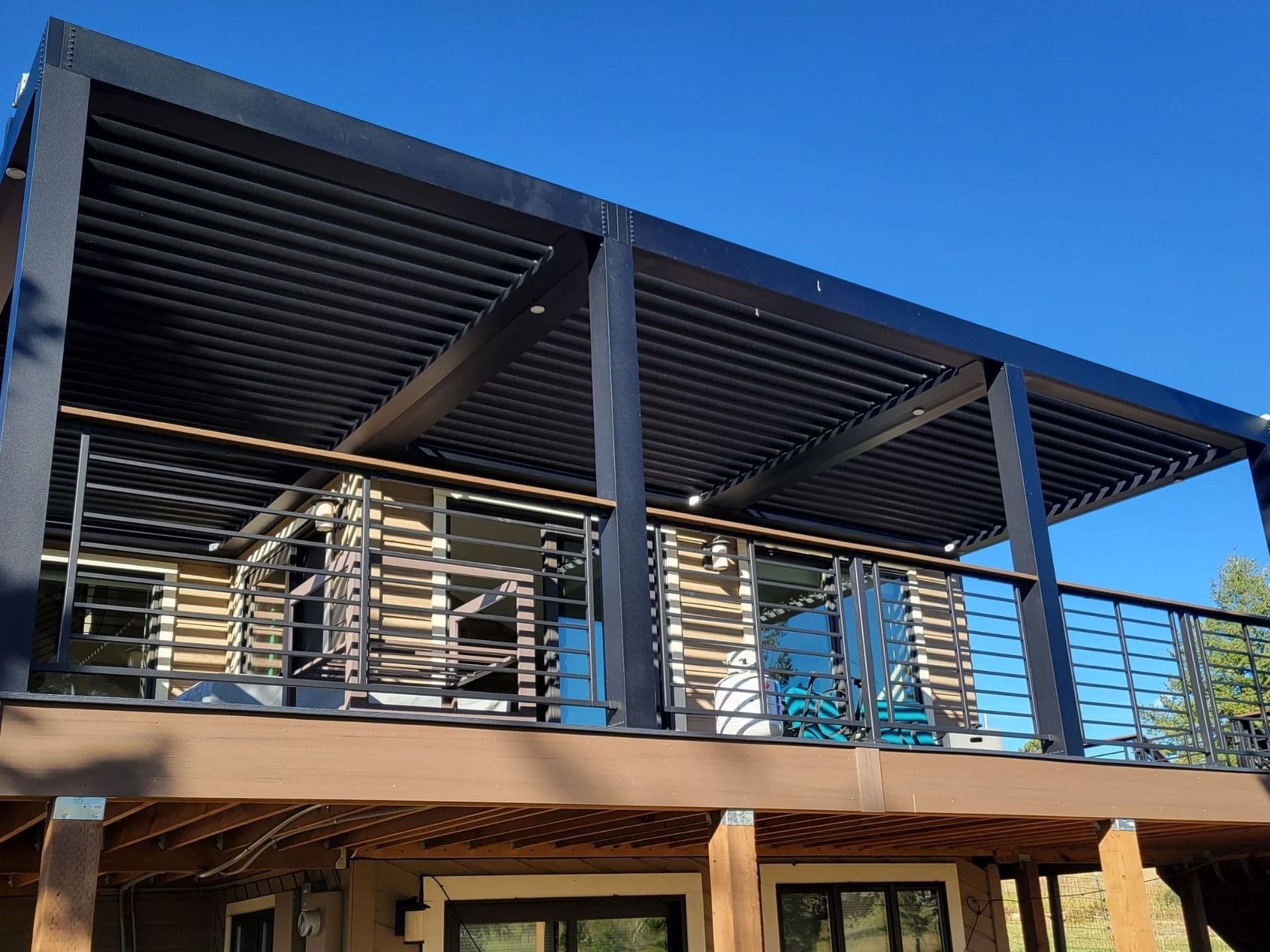 StruXure of the Rockies installed two pergolas for this homeowner in the foothills west of Boulder, CO, at an elevation of about 10,000 feet. The property faced extreme weather conditions characterized by high winds and heavy snow loads. The homeowner wanted two different pergola systems: one on a small, elevated front deck and another on a large, elevated wrap-around deck. The first pergola was installed over a small (10 x 12) front deck, providing a covered space for the homeowner to enjoy morning coffee while taking in the stunning mountain views. This deck, elevated 5-6 feet off the ground, was designed to offer protection from the elements without obstructing the natural beauty of the surroundings. The second pergola was part of a larger project involving a complete teardown and reconstruction of the old wrap-around deck. Working closely with Legacy Spaces, a renowned deck builder, StruXure of the Rockies ensured that the new deck was perfectly sized to accommodate the pergola. The deck design and creation involved several steps, including tearing down the old deck, designing the new one, constructing it, and installing the pergola and railings. This collaborative effort resulted in an integrated and seamless outdoor living space. The second pergola, covering approximately 450 square feet, featured three zones to cater to a large main section of the wrap-around deck. The bronze-colored Pivot 6 pergola was equipped with lighting, though no fans were installed due to the high wind conditions in the area.